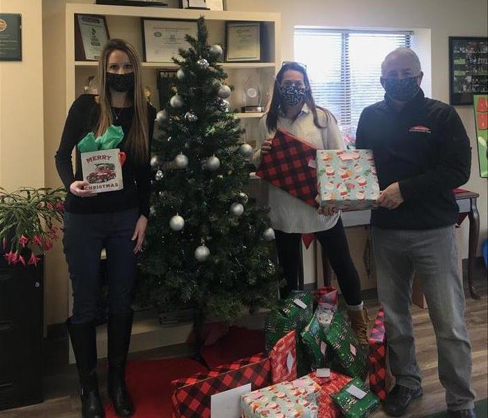 2 females and 1 male standing near Christmas tree holding gifts